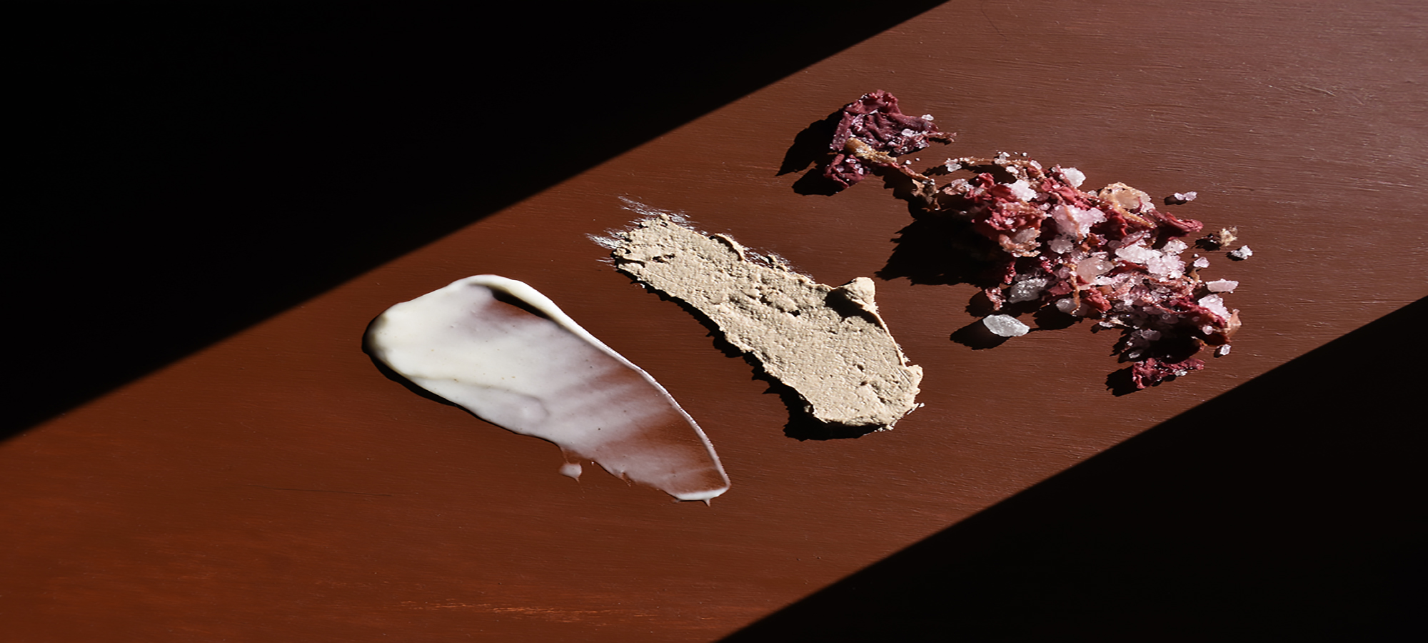 Three different types of skin care products smeared across a red/brown background with a slice of light coming in at an angle from the top right hand corner creating shadow on either side of the product